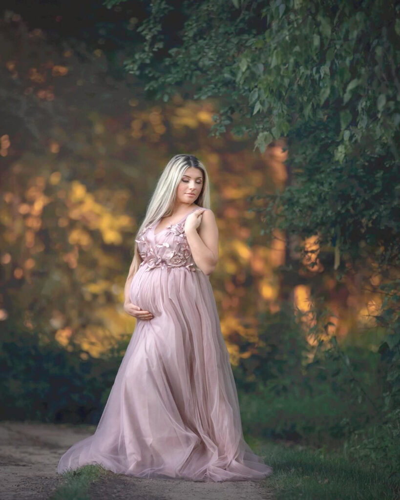 Pregnant woman in a flowing blush gown standing in a dreamy outdoor setting at golden hour, cradling her baby bump. A perfect fine art maternity portrait illustrating when to book a maternity session."