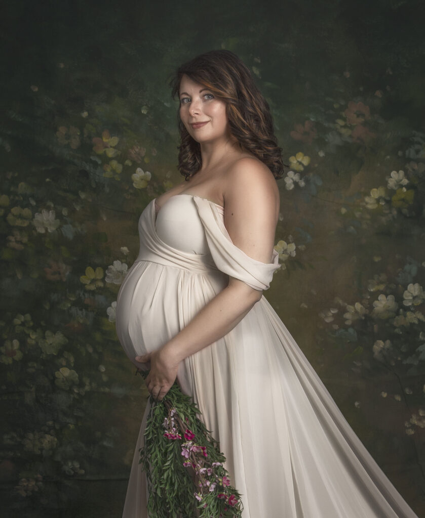 Pregnant woman in an elegant off-shoulder gown cradling her baby bump, set against a fine art floral backdrop. A perfect example of when to book a maternity session with Sonia Gourlie Fine Art Photography.