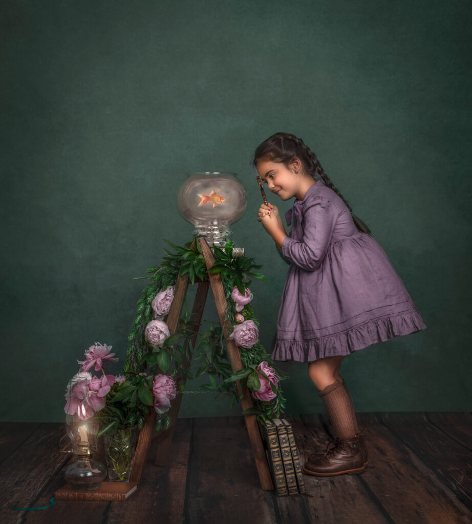 A young girl in a studio setting, curiously observing her goldfish through a magnifying glass, captured by Sonia Gourlie Fine Art Photography. The serene studio atmosphere adds a sense of wonder and exploration to the moment between the girl and her pet fish. Perfect for pet photoshoot timings and capturing precious moments with your pets.”
