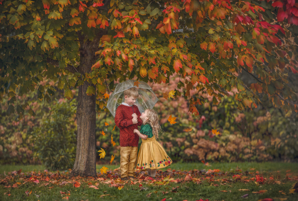 Two young children playing under a colorful umbrella as autumn leaves fall around them during a cozy fall photography session.