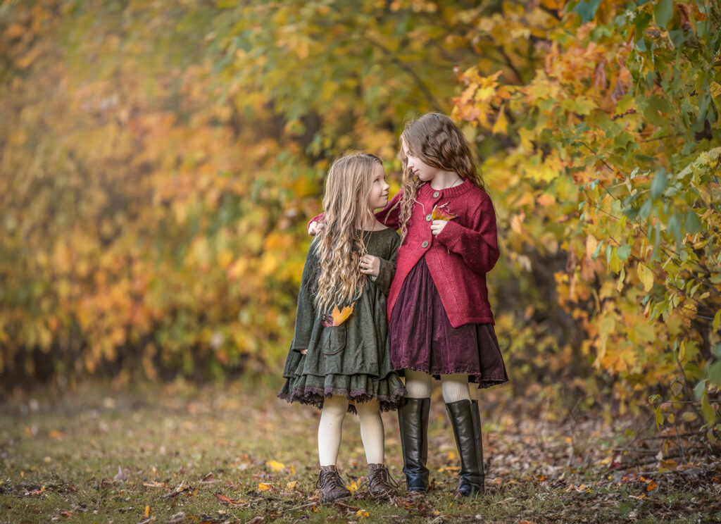 Two sisters sharing a sweet, candid moment while looking at each other surrounded by vibrant fall leaves